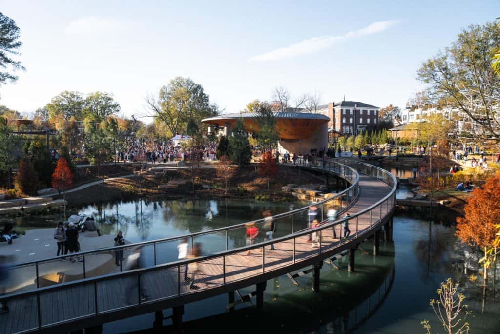 A vibrant sunny day at Downtown Cary Park in North Carolina, featuring a curved wooden pedestrian bridge over a pond, a modern pavilion, and people enjoying the outdoors.