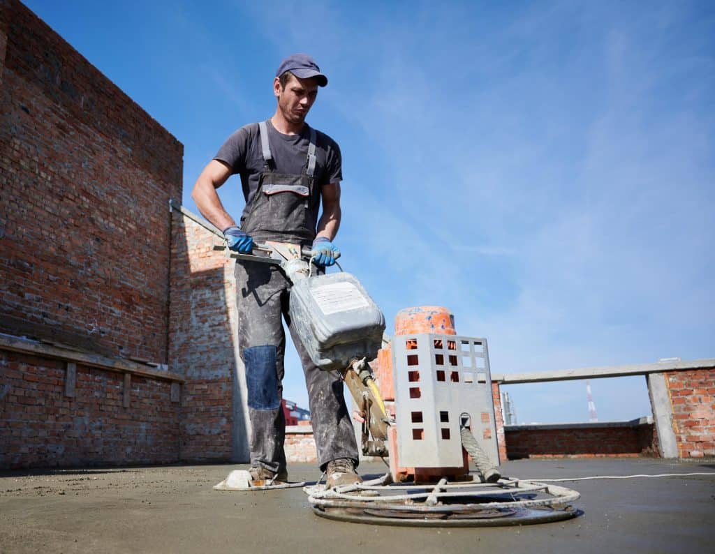 A construction worker in overalls using a heavy-duty power trowel machine to smooth wet concrete on an industrial building site.