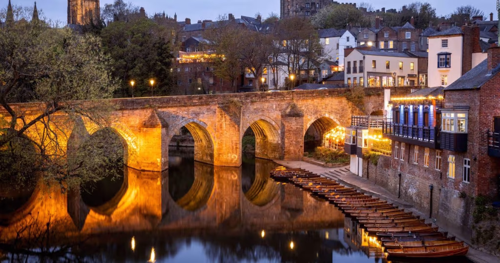 An illuminated historic stone arched bridge crossing a river at dusk in a European city.