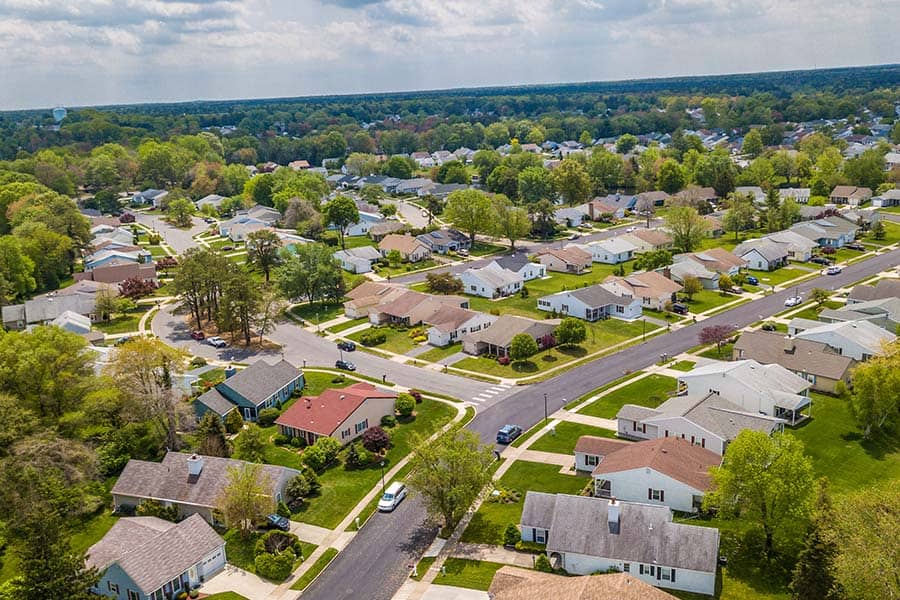 An aerial view of a lush, green suburban neighborhood with houses, manicured lawns, and tree-lined streets.