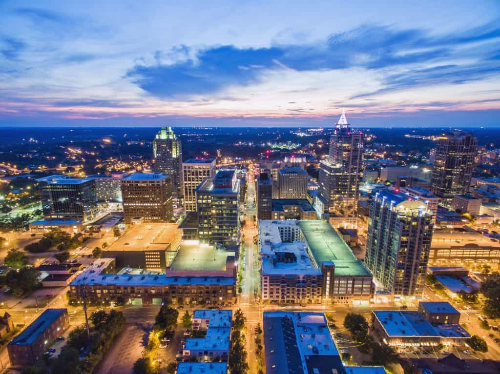 An aerial nighttime view of the illuminated downtown Raleigh skyline under a twilight sky.